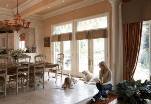 A mother, child, and dog sit in their dining room next to a large pair of garden doors. 