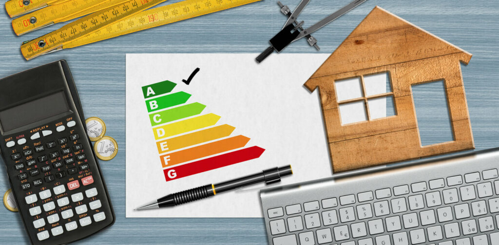 House energy efficiency rating. Small wooden model house on desk with energy efficiency graph, calculator, folding ruler, drawing compass, pencil, and a computer keyboard.